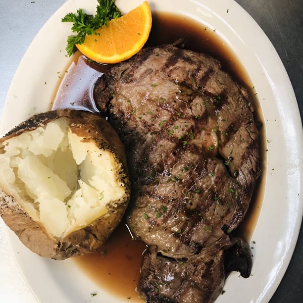 Grilled steak with a baked potato, garnished with parsley and an orange slice, on a white plate.