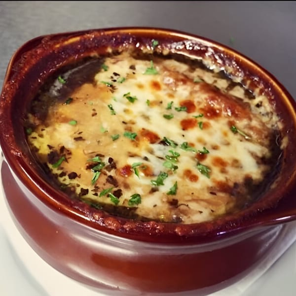 A rich, bubbly French onion soup in a brown crock, topped with melted cheese and herbs.