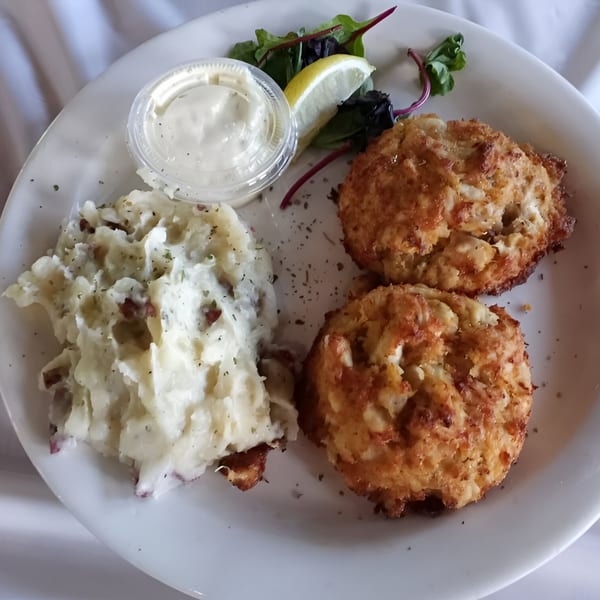 Two golden-brown crab cakes with mashed potatoes, tartar sauce, lemon, and greens on a white plate.