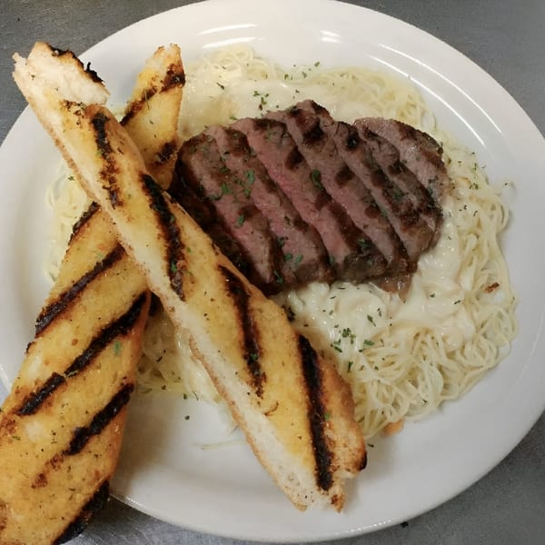 Grilled steak slices over creamy pasta with two pieces of grilled garlic bread on a white plate.